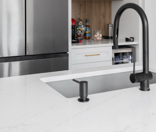 Évier encastré avec robinet noir moderne sur un comptoir en quartz blanc, accompagné d’armoires de cuisine blanches et d’un espace de rangement en bois intégré.