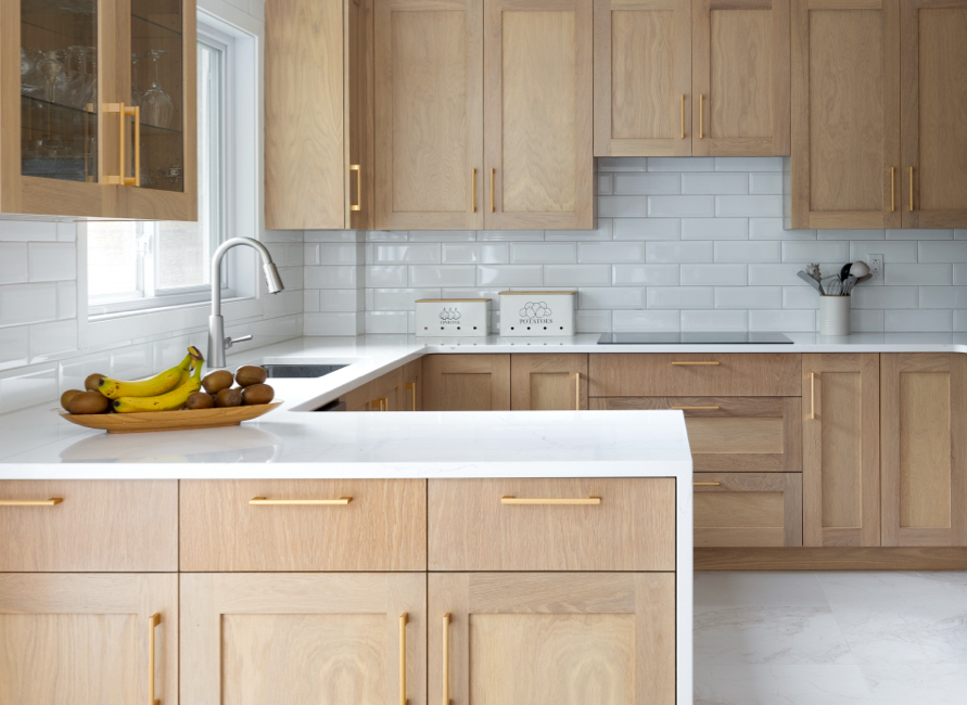Cuisine chaleureuse avec armoires en bois clair, poignées dorées et comptoirs en quartz blanc, sublimée par un dosseret en carrelage métro pour un style intemporel.