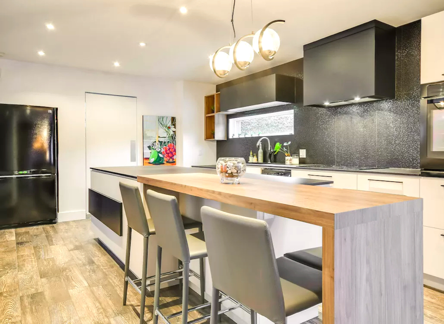 Comptoir de cuisine en bois massif avec îlot central, combinant élégance naturelle et design contemporain. Tabourets confortables, éclairage suspendu moderne et finitions noires apportent une touche sophistiquée.