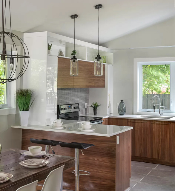 Modern kitchen with a central wood island and a white countertop, illuminated by designer pendant lights. Large windows provide natural light and a view of a lush green garden.