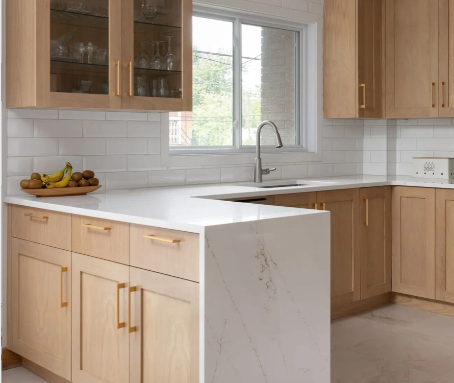 Modern light wood kitchen with a white quartz countertop and a subway tile backsplash. A sink positioned under the window brings in natural light, enhanced by gold handles for an elegant touch.