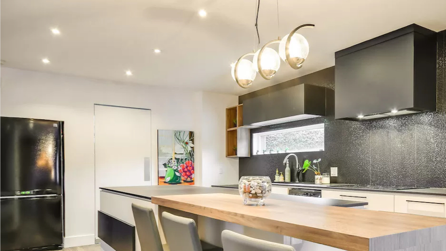 Modern kitchen with a central wood island and stylish spherical pendant lights. Elegant black cabinetry, a textured backsplash, and matching appliances create a contemporary and warm ambiance.